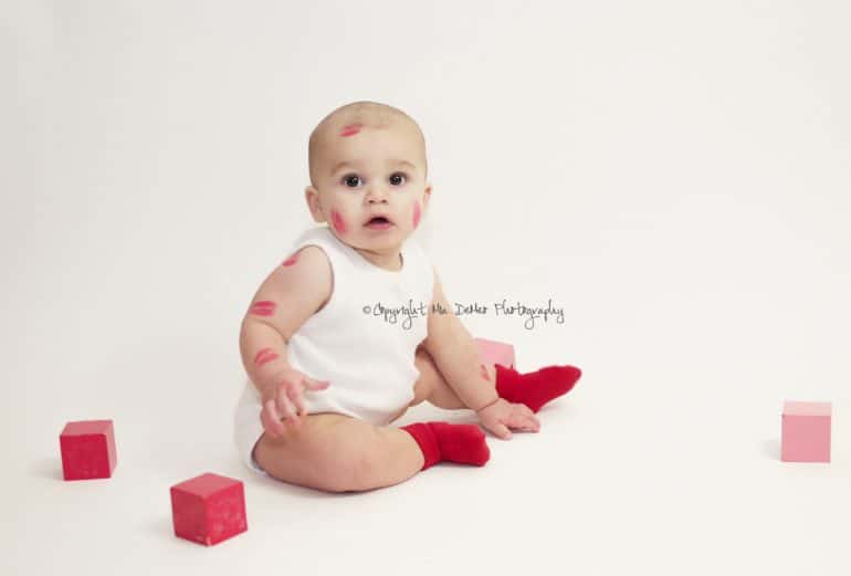 Baby sitting on floor with pink on red blocks and kiss marks on their face and arms - Baby's First Valentine's Day Photo shoot