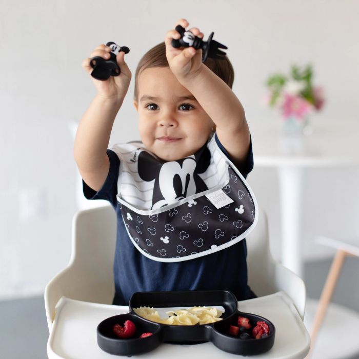 Toddler sitting in a highchair wearing Mickey Mouse bib, eating off Mickey Mouse plate, playing with Mickey Mouse utensils.