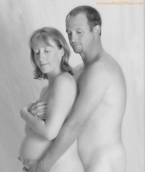 Fully nude couple spoons while she covers her breasts with her hands and he cradles her bump.