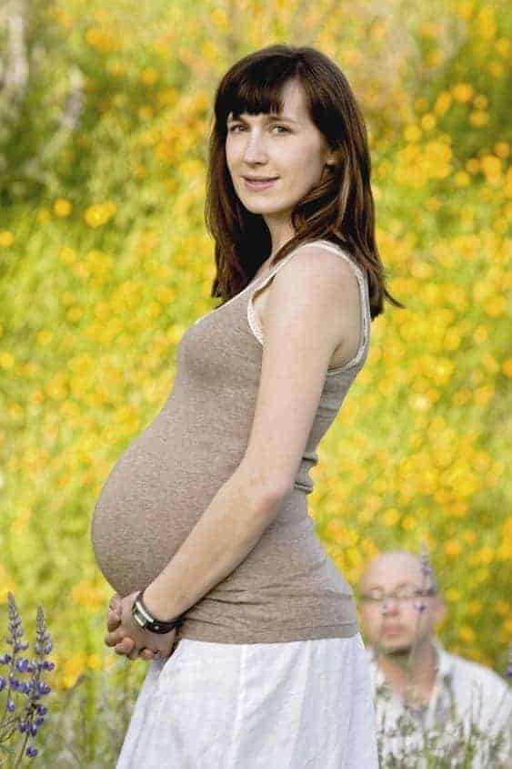 Pregnant woman stands in field of wildflowers while bald man crouches in the background