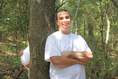 Photo of a man standing cross-armed in a forest next to a tree where nothing but a baby bump is shown peeking out from behind.