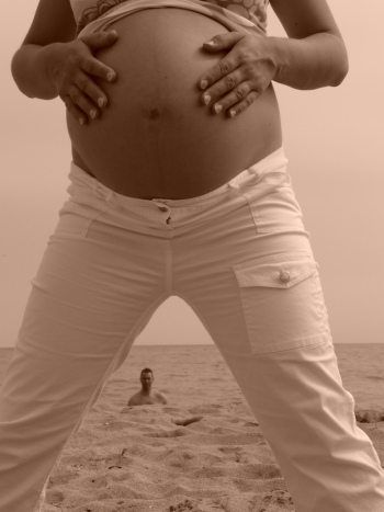 Black and white image of woman standing, wide-legged, on a beach while man peers from a distance between her legs.