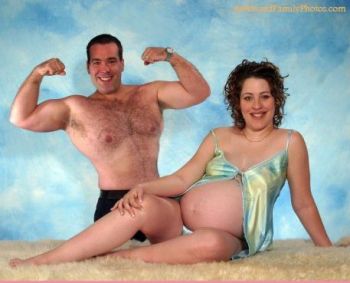 Couple faces the camera, he, standing behind his pregnant partner, topless and flexing, she sitting and smiling while her shirt exposes her bare bump.