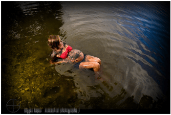 Woman lays in muddy river with red top, black shorts, and mud smeared all over her belly.