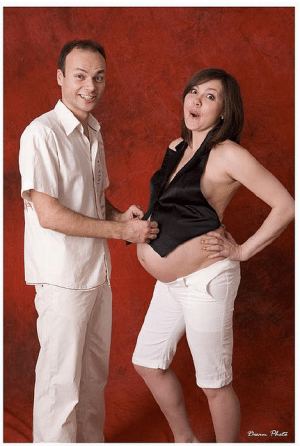 Couple stands facing each other smiling while he holds onto tiny black vest she's wearing as a top.