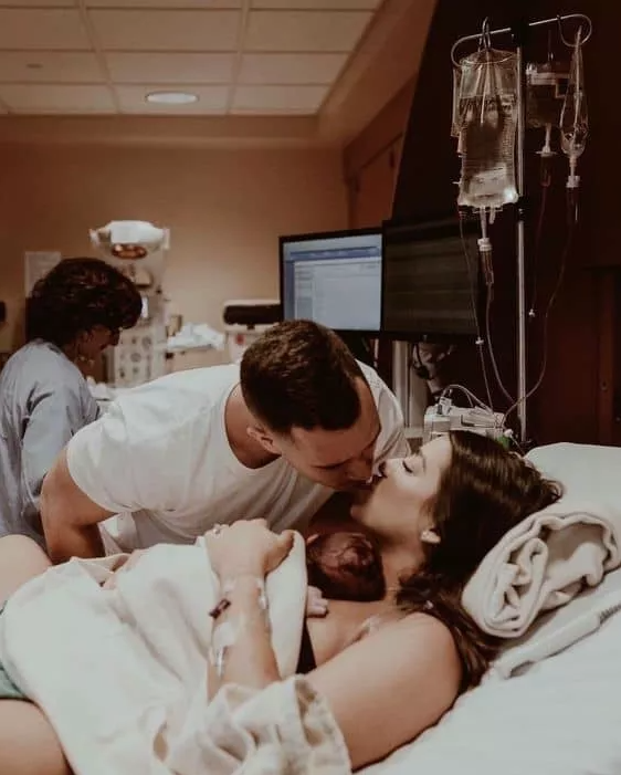Color image of dad leaning down to kiss mom while she lays with newborn sleeping on her chest.