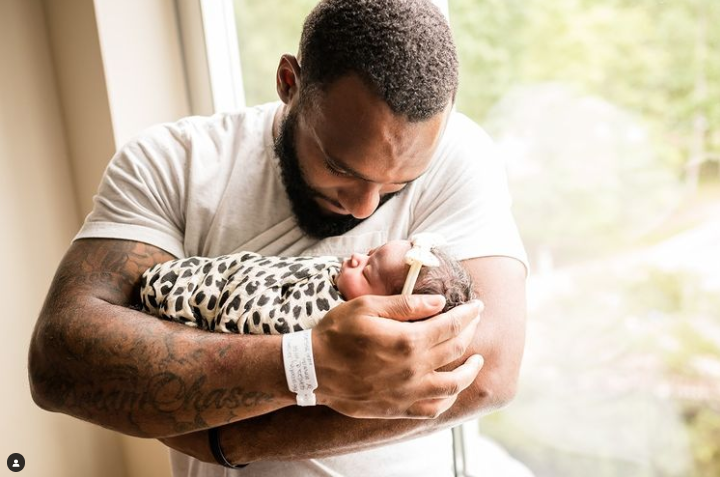 Color photo of dad holding newborn swaddled in cheetah print in front of window.