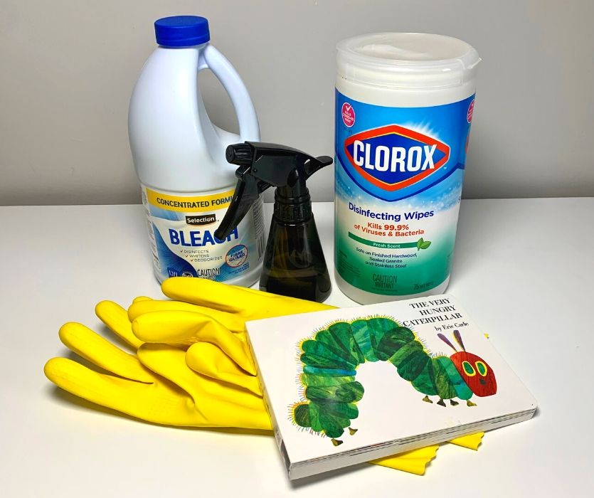container of bleach, spray bottle, board book, disinfecting wipes and yellow rubber gloves