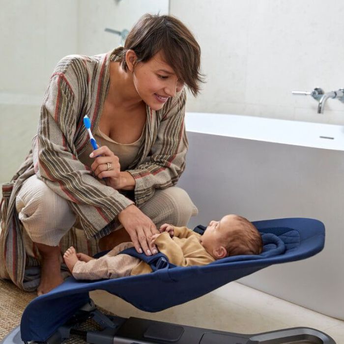 mom with toothbrush looks at baby in bouncer in bathroom